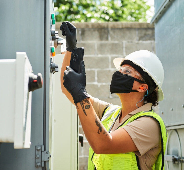 A worker in a hard hat and safety vest uses her phone in front of outdoor equipment.
